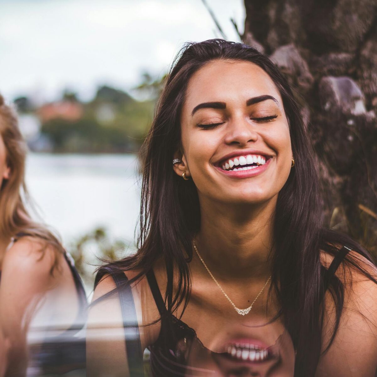 Two women laughing together outdoors near a tree, sharing a relaxed moment with natural smiles