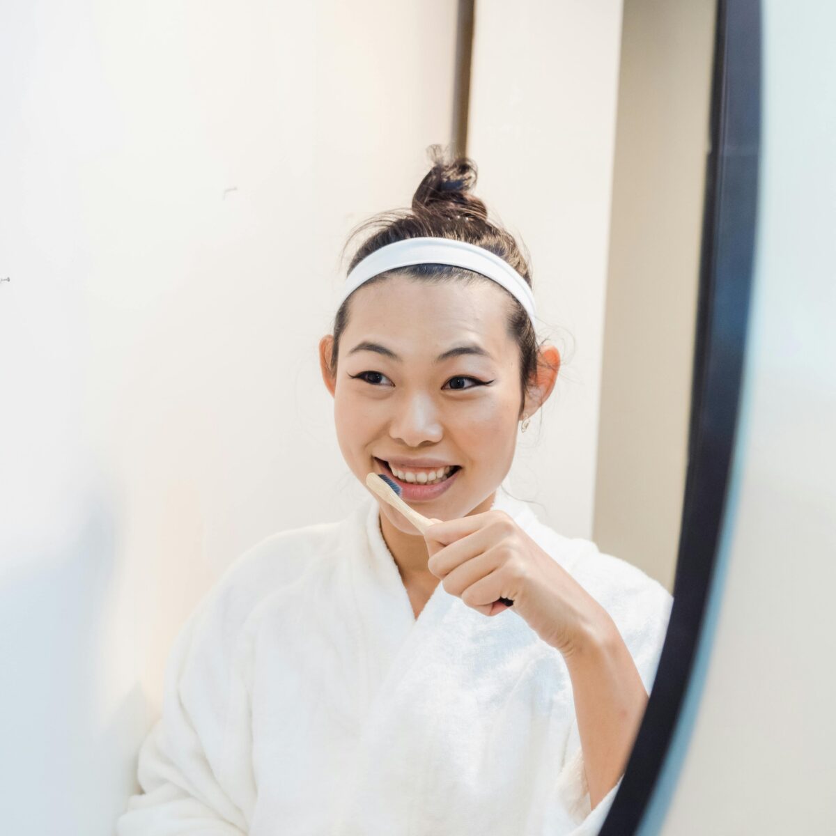 Woman in a robe brushing her teeth while smiling at her reflection in a bathroom mirror