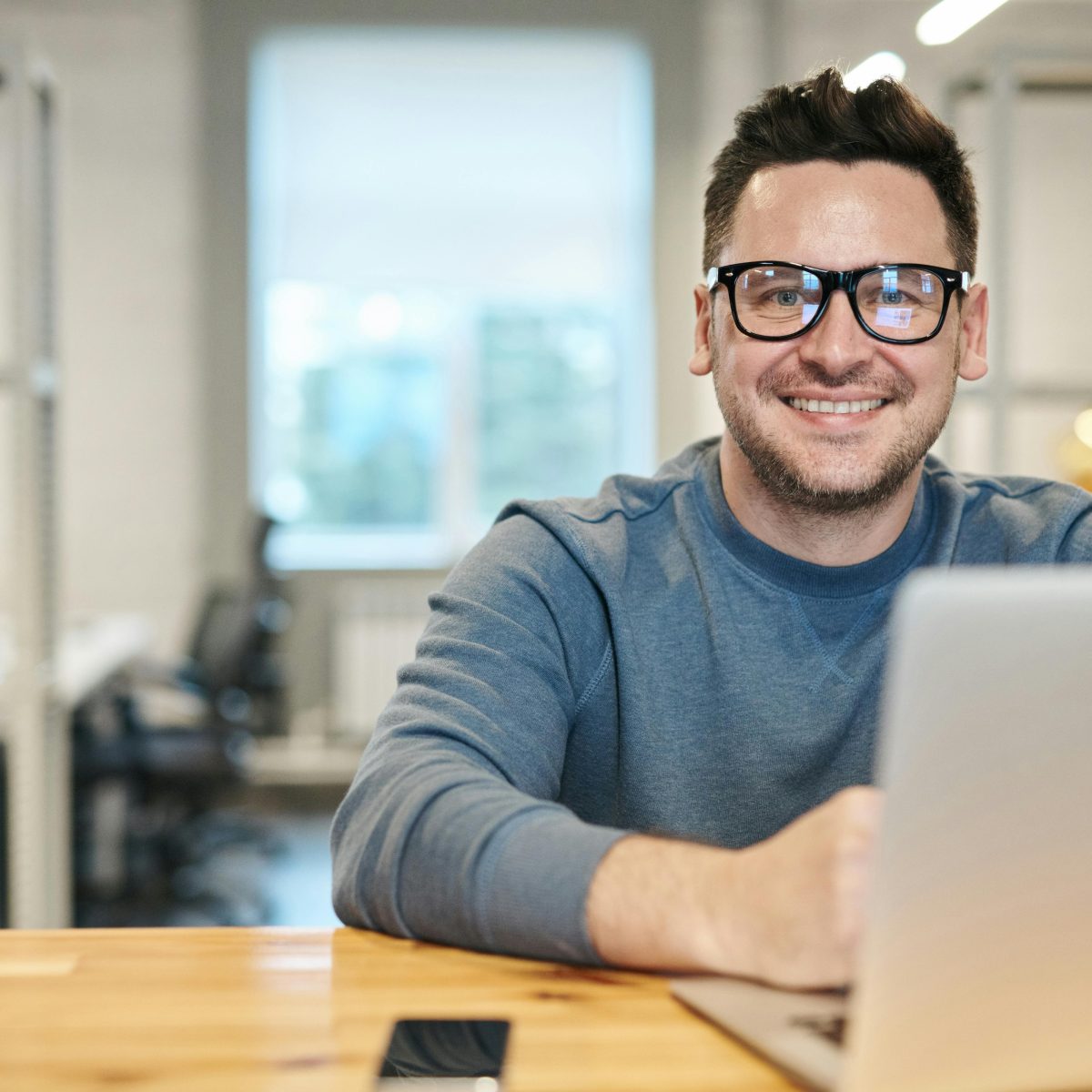 Man smiling at a laptop in a bright office setting, wearing glasses and casual clothing
