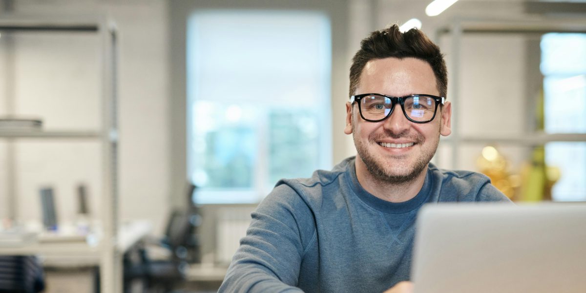 Man smiling at a laptop in a bright office setting, wearing glasses and casual clothing