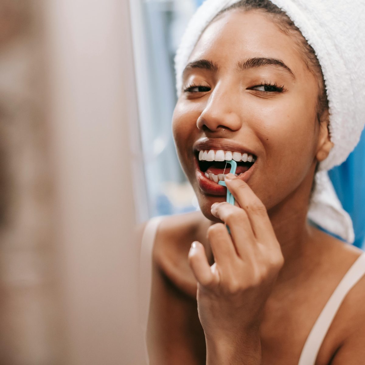Woman flossing her teeth while smiling in a bathroom mirror after getting ready