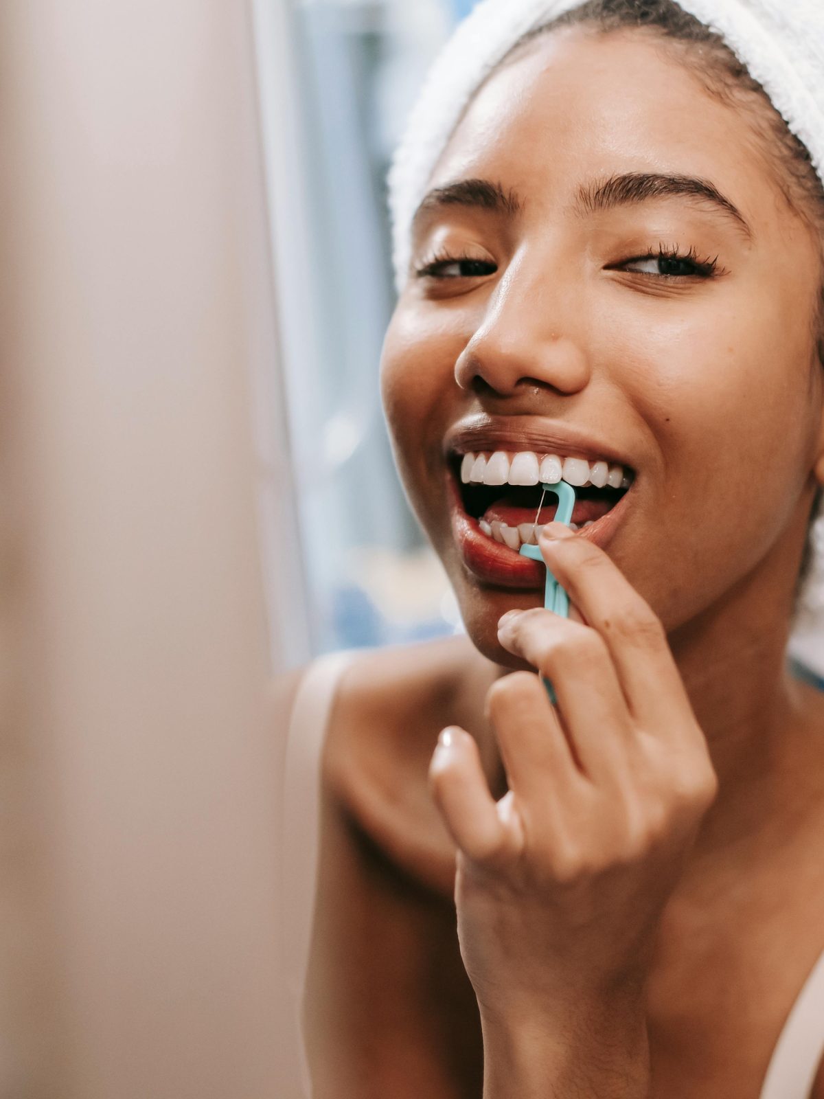Woman flossing her teeth while smiling in a bathroom mirror after getting ready
