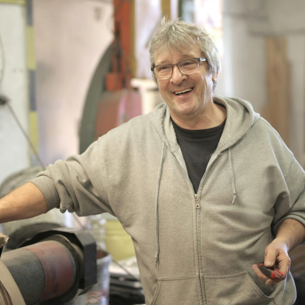 Older man smiling in a workshop, wearing a hoodie and standing beside industrial equipment