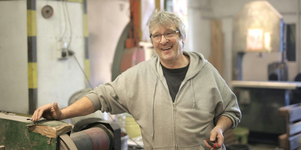 Older man smiling in a workshop, wearing a hoodie and standing beside industrial equipment