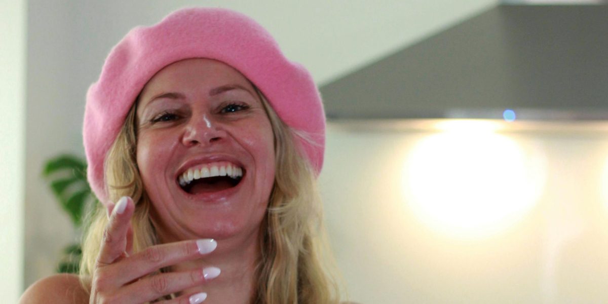 Woman laughing in a kitchen with a bright, natural smile, wearing a pink beret and apron