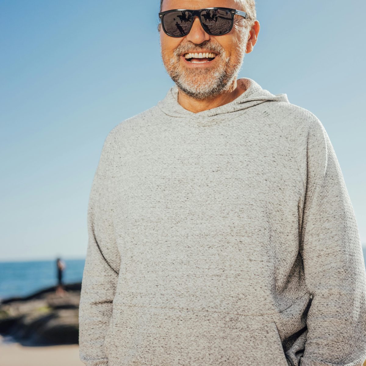 Older man wearing sunglasses and a hoodie, smiling naturally outdoors under a clear blue sky