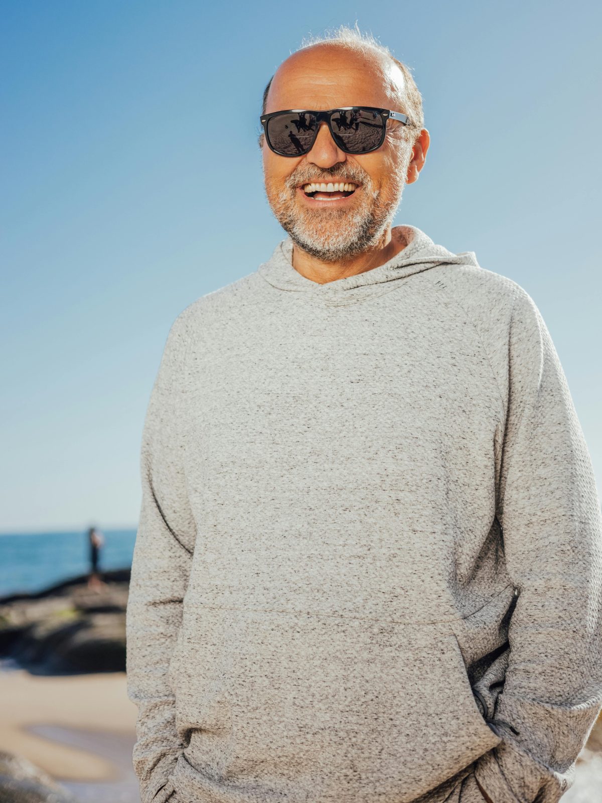 Older man wearing sunglasses and a hoodie, smiling naturally outdoors under a clear blue sky