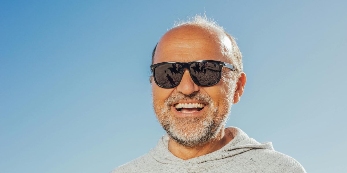 Older man wearing sunglasses and a hoodie, smiling naturally outdoors under a clear blue sky
