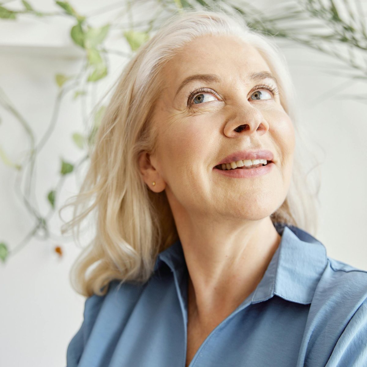 Older woman with light hair smiling softly indoors, looking upward in natural daylight with plants in the background