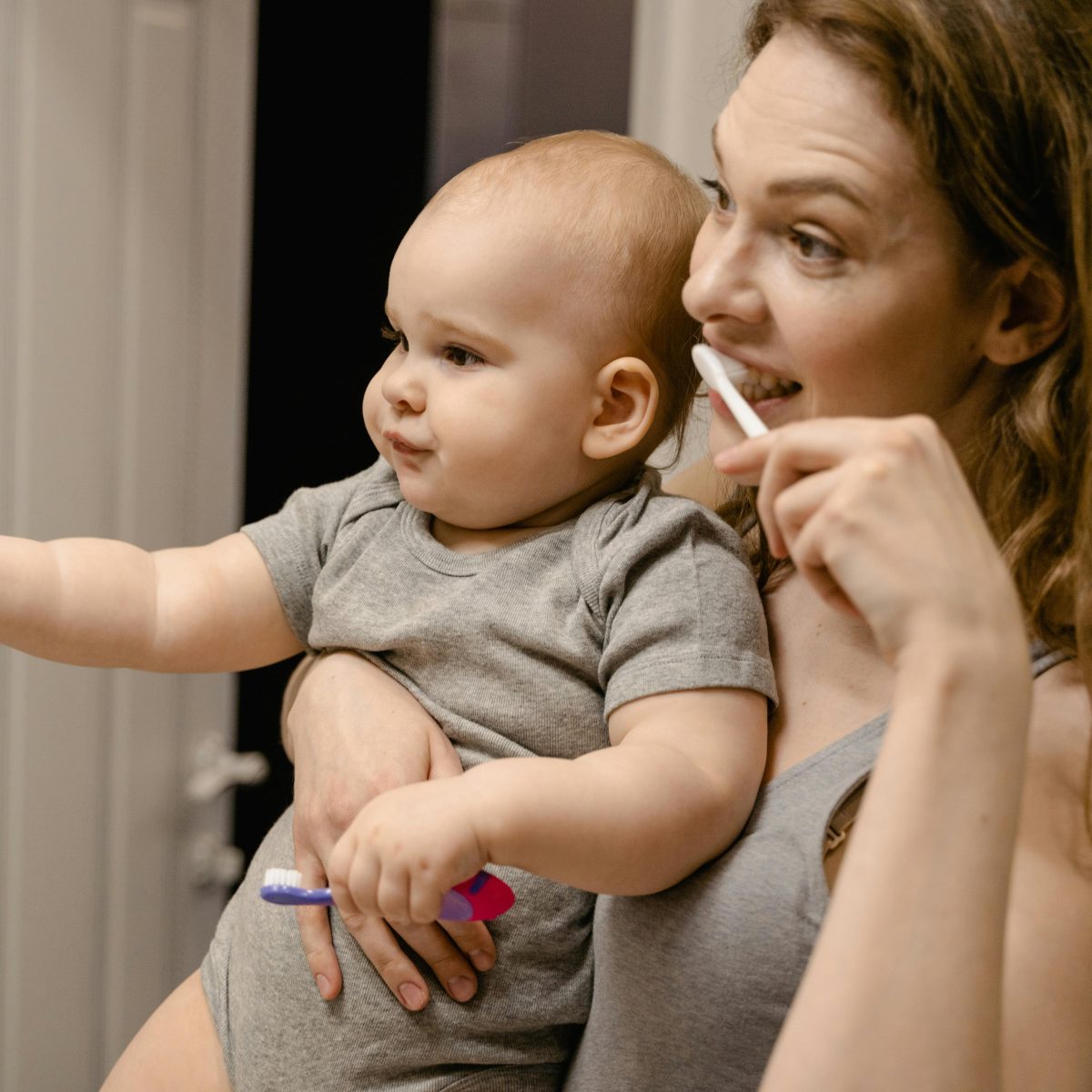 Mother holding a baby while brushing her teeth in a bathroom, sharing a gentle everyday moment