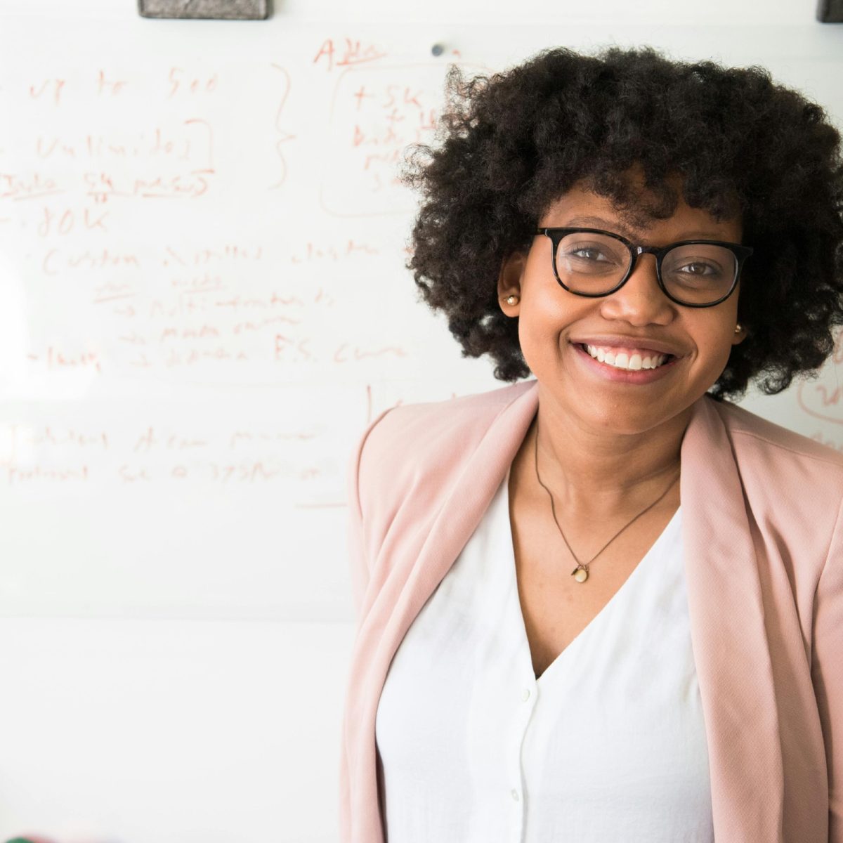 Woman smiling confidently in a professional setting, wearing glasses and standing in front of a whiteboard
