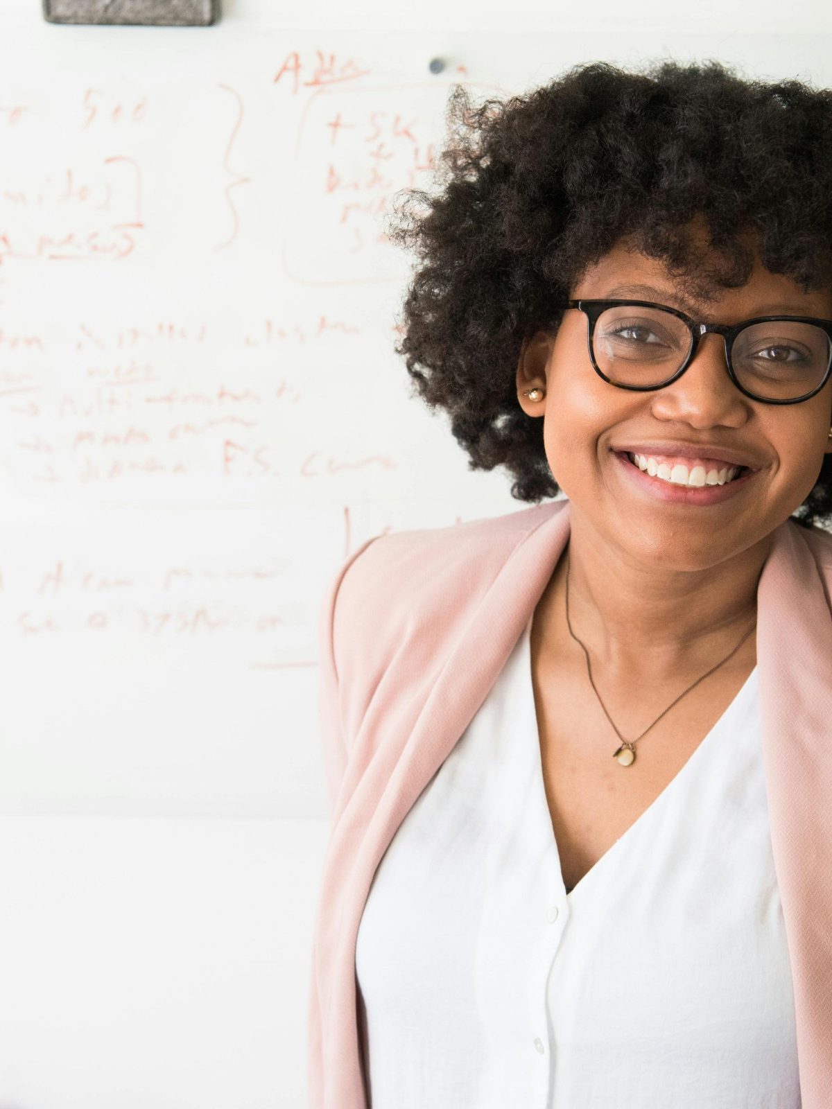 Woman smiling confidently in a professional setting, wearing glasses and standing in front of a whiteboard