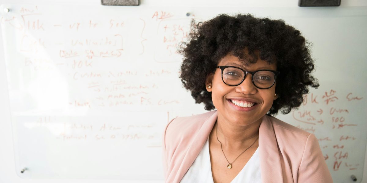 Woman smiling confidently in a professional setting, wearing glasses and standing in front of a whiteboard