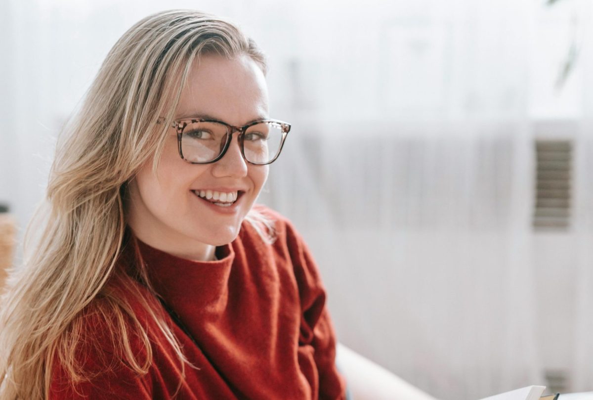 Woman wearing glasses smiling while reading a book in a cozy indoor setting
