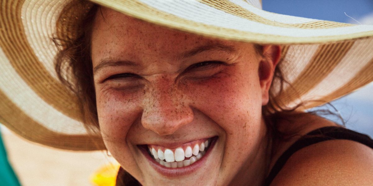Woman wearing a sun hat smiling broadly outdoors, showing natural teeth and freckles